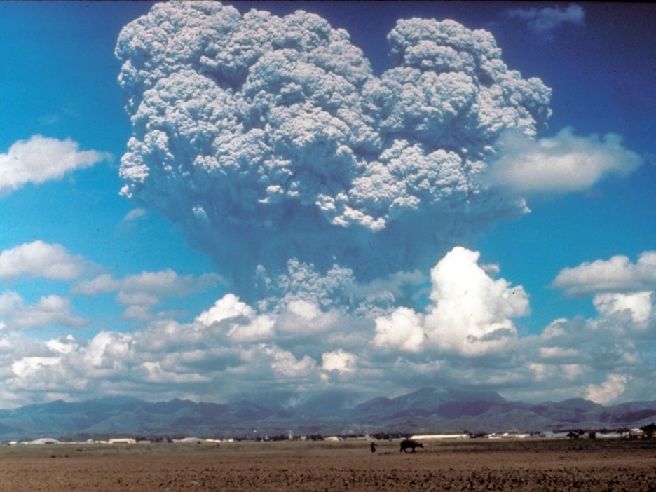 Volcanic eruption with a massive ash cloud against a blue sky over barren land and distant mountains.